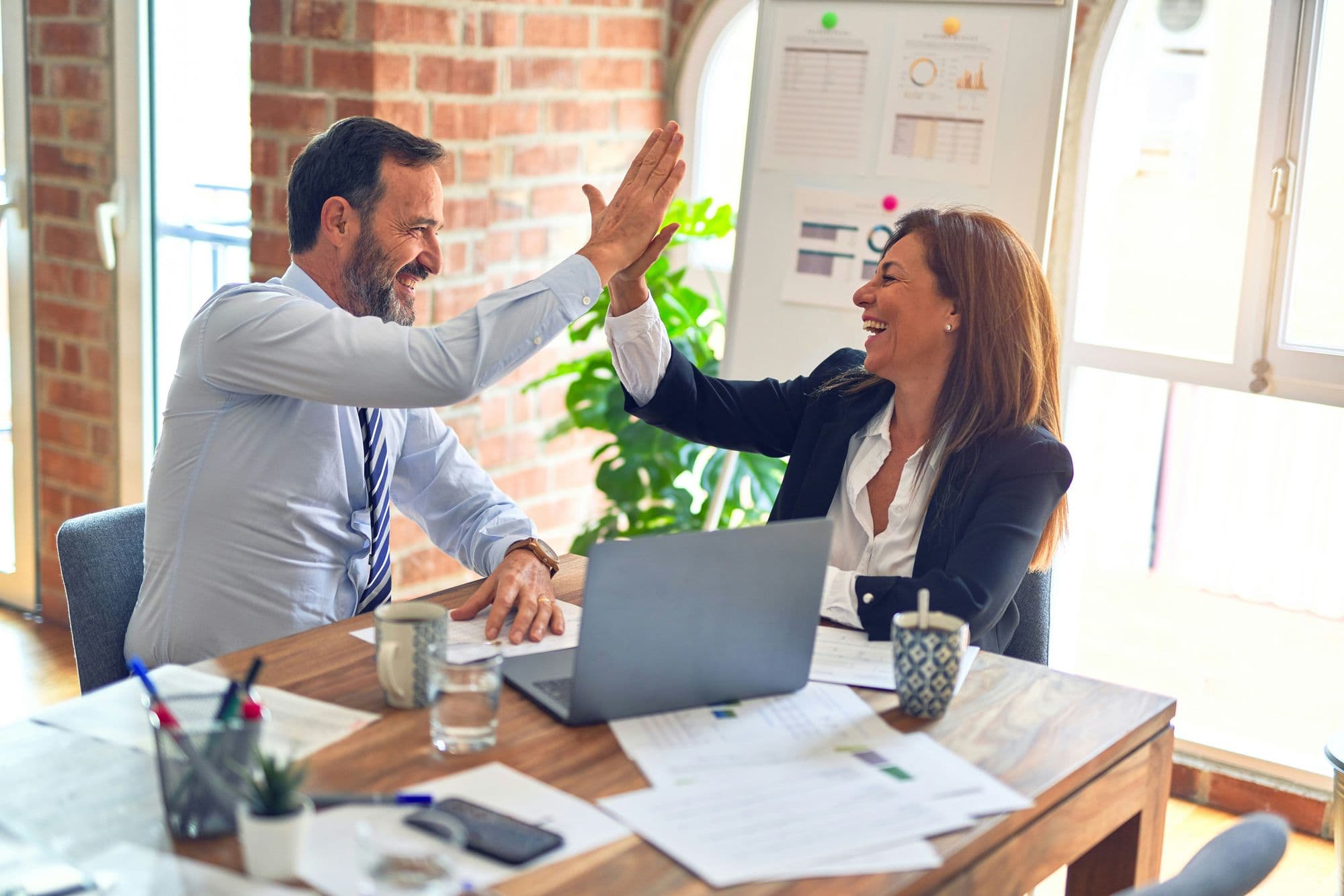 Two business colleagues high-fiving across a desk in a brick-walled office