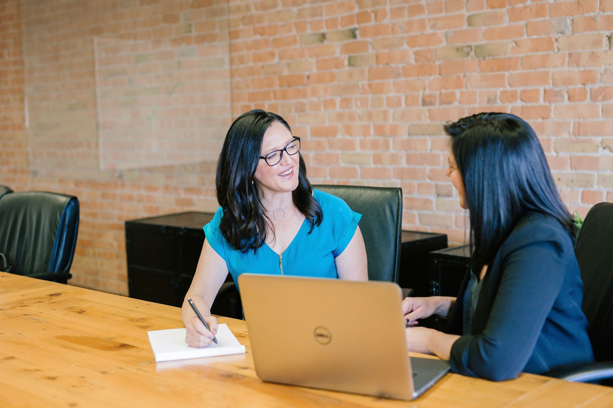 Two businesswomen discussing notes over a laptop in a brick-walled meeting room